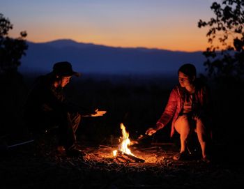 Man and woman sitting by bonfire against sky during sunset