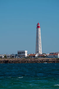 Lighthouse by sea against buildings against clear blue sky