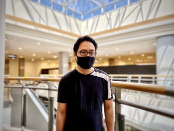 Portrait of young man in eyeglasses and face mask standing against architecture and skylight.