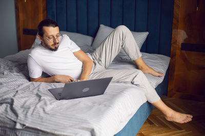 Man in a white t-shirt, a freelancer with glasses, sits on the bed at home with a laptop and works