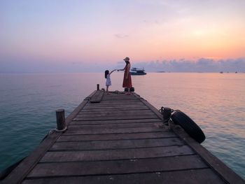 Side view of mother with daughter standing on pier over sea against sky during sunset