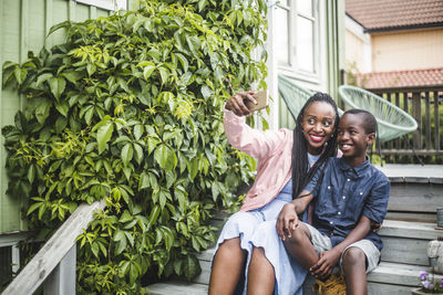 Mother taking selfie through smart phone while sitting by plants on steps