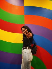 Portrait of young woman showing peace sign while standing against colorful wall