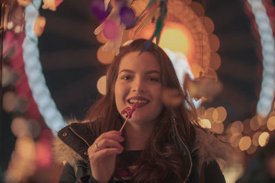 Portrait of young woman eating food while standing against illuminated lights at night