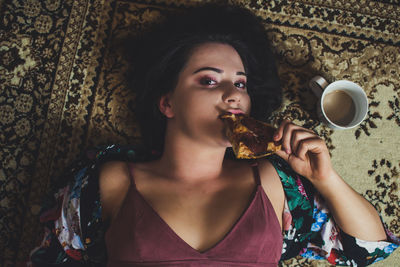 High angle portrait of young woman eating pizza while lying on carpet at home
