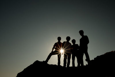 Sun streaming through silhouette friends standing on cliff against clear sky