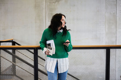 Young student with books and mobile phone looking away while standing at railing against wall in university