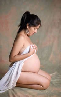 Side view of naked pregnant woman covering breast while kneeling against backdrop