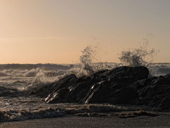 Waves splashing on rocks at beach against clear sky during sunset