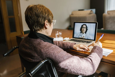 Senior woman sitting on wheelchair writing in book while video consultation at home during covid-19