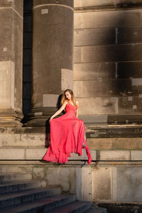 Full length of young woman in pink evening gown sitting against columns