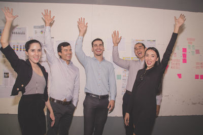 Portrait of happy colleagues with arms raised standing against wall in office