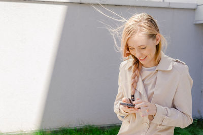 Smiling young woman using smart phone against wall