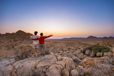 Rear view of men standing on rock against sky during sunset