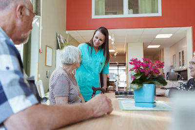 Smiling caregiver assisting senior woman sitting with friends at nursing home