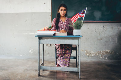 Portrait of young teacher sitting at table against blackboard in classroom