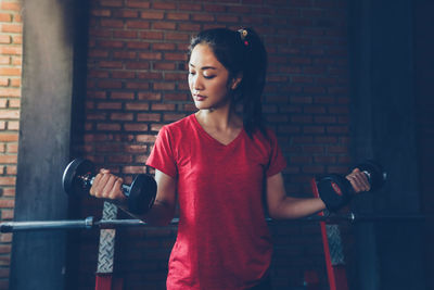 Young woman lifting dumbbells while standing against brick wall