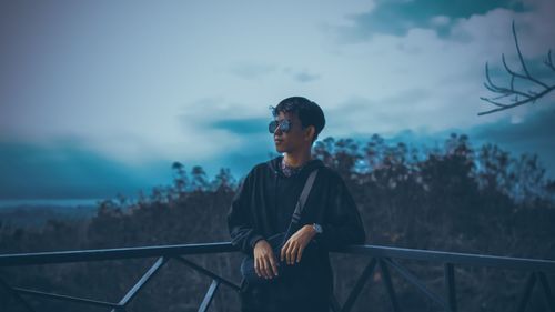 Teenage boy wearing sunglasses standing by railing against sky at dusk