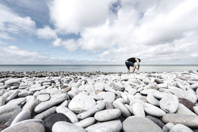 Man collecting pebbles on shore against sky