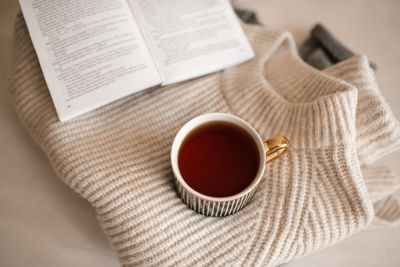 Cup of coffee stay on knitted textile sweater in bed with paper open book close up. winter cozy.