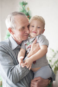 Sentimental old man holding small child in his arms  in front of decorated christmas tree