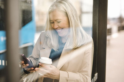 Happy businesswoman using smart phone holding disposable cup while leaning on glass at bus stop