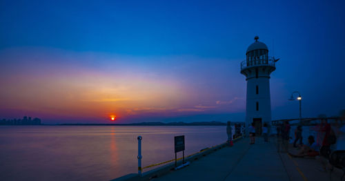 Lighthouse amidst sea and buildings against sky during sunset