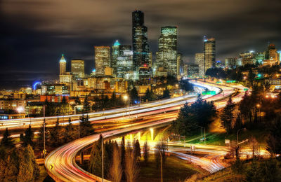 Light trails on road by illuminated buildings against sky at night