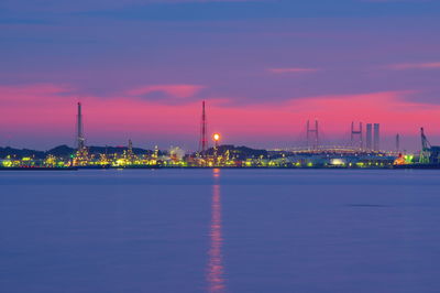 Illuminated city by sea against sky at sunset