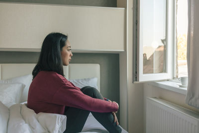 Side view of young woman looking through window while sitting on bed at home