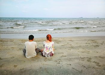 Rear view of friends sitting on beach against sky