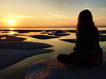 Woman sitting on rock at sea shore against sky during sunset