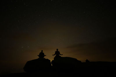 Silhouette people sitting on rock against sky at night