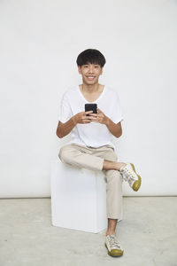 Portrait of smiling man holding smart phone while standing against wall