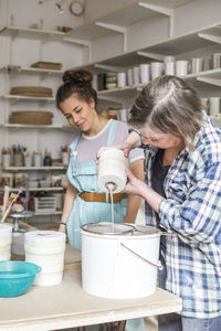 Young employee looking at mature female potter pouring clay from vase in strainer on bucket at workshop