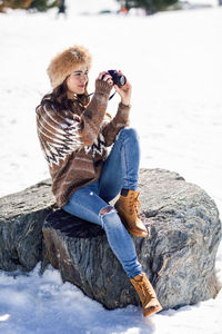 Full length of young woman photographing while sitting on rock during winter