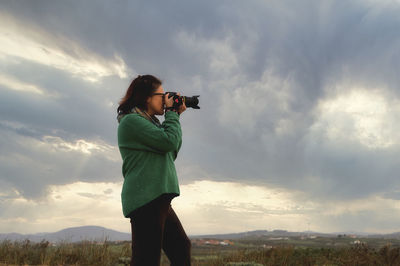 Woman photographing with camera while standing on mountain against sky during sunset