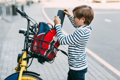 Boy in a striped sweater puts a tablet in a backpack that hangs on the handlebars of a bicycle