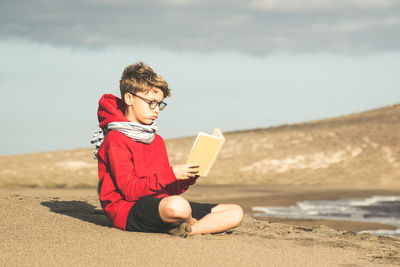 Boy reading book while sitting at beach against sky