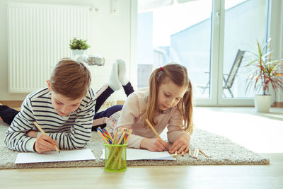 Siblings writing on paper while lying at home