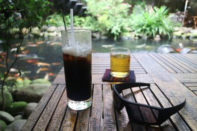 Close-up of drinks and sunglasses on wooden table against lake in forest