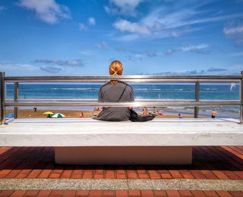 Woman sitting on bench at beach against sky