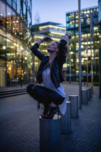 Young woman crouching on bollard in city at night