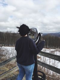 Rear view of woman standing by coin-operated binoculars against cloudy sky
