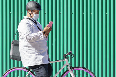 Mature man sitting on bicycle using smart phone by green metal wall