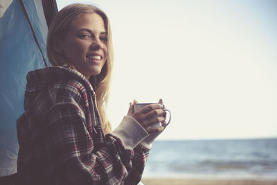 Portrait of smiling young woman drinking coffee while standing at beach against sky
