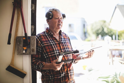 Thoughtful senior man holding clamp and digital tablet at workshop doorway