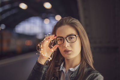Close-up portrait of young woman holding illuminated string lights while standing at railroad station platform at night