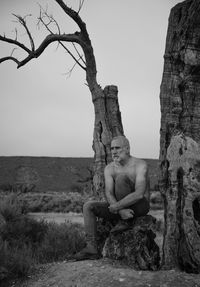 Monochrome of shirtless man sitting on abare olive tree against sky