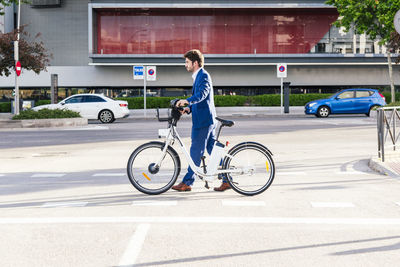 Side view of trendy unshaven male office worker in formal wear strolling on street with electric bicycle